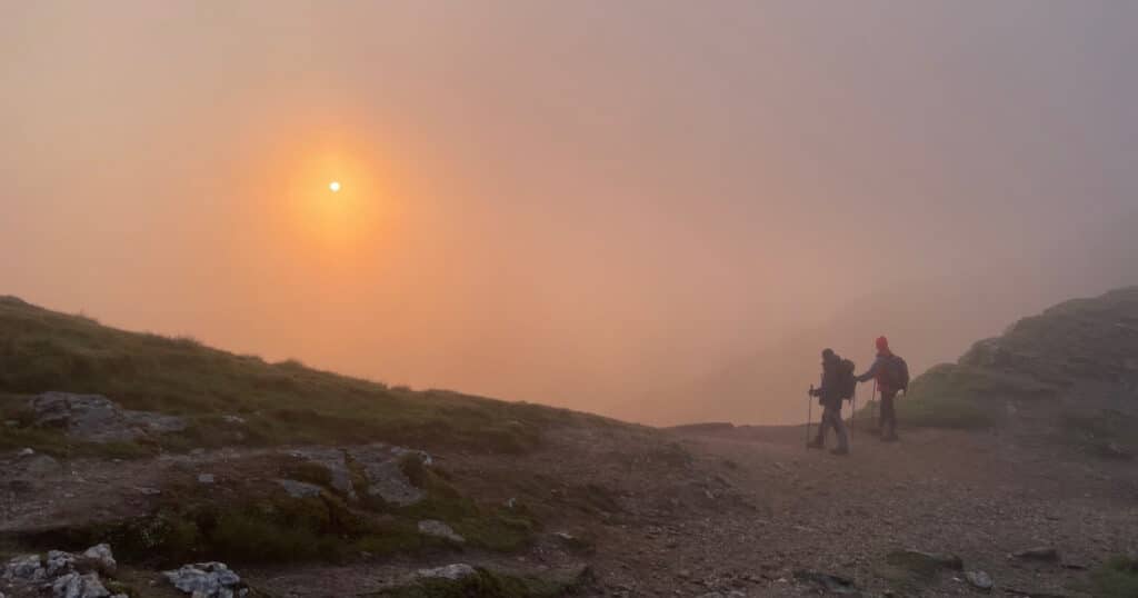 2 figures hillwalking on top of mountain with sun rising through fog