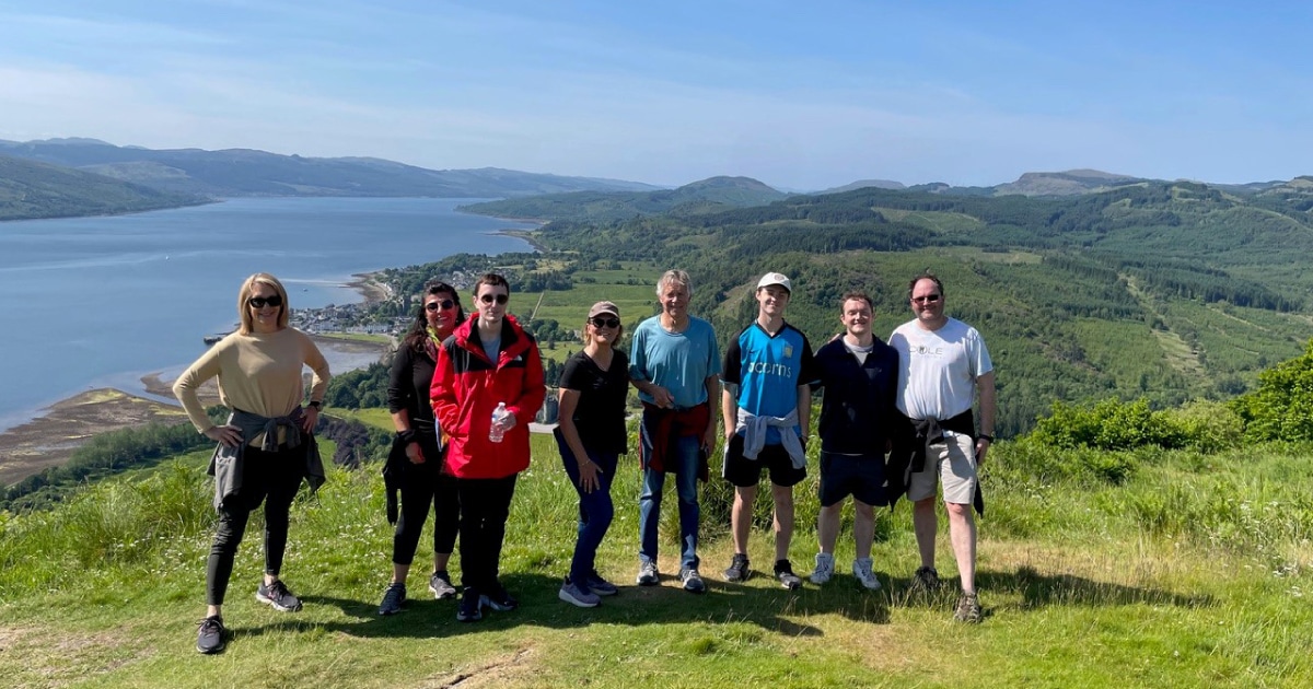 Group of 8 walkers on top of a hill in sunshine with view over loch and hills in background