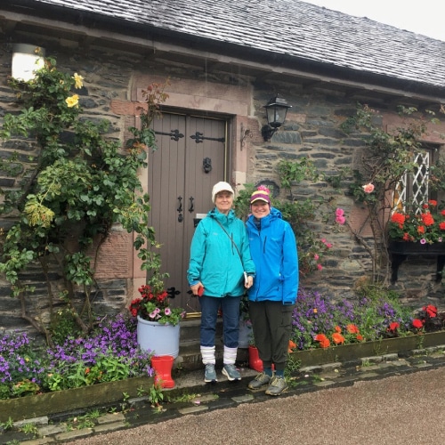 2 people in waterproof clothes standing on the doorstep of a stone cottage with flowers around the building